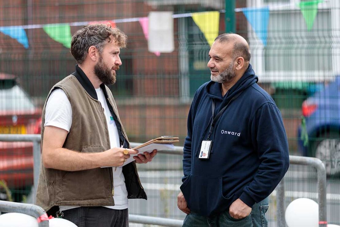 Two men, one with a lanyard and one with a clipboard, stand in conversation outside. There are parked cars and colourful bunting behind them.