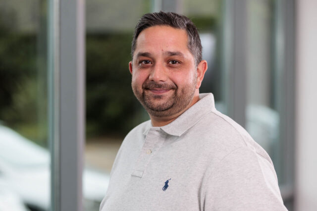 Photograph of a man wearing a light grey polo shirt with a small dark blue logo on the left chest, standing indoors near large windows. Background shows blurred greenery and parts of a white vehicle outside.