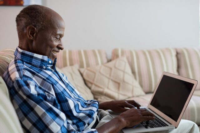 Photograph of an elderly man sitting on a striped couch using a laptop. The scene highlights a casual home environment with neutral-toned cushions and the man wearing a blue plaid shirt.