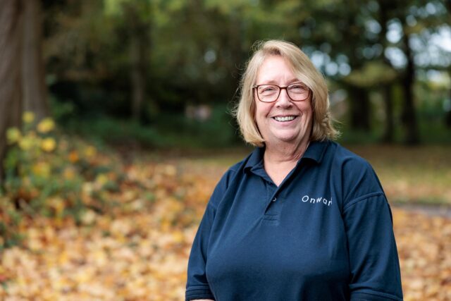 Photograph of a person wearing a navy blue polo shirt with the Onward logo standing outdoors in a park with autumn leaves on ground and trees in background.