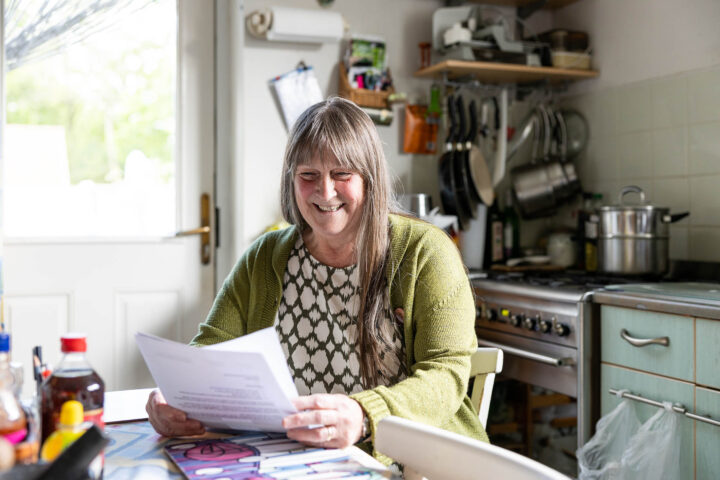 Photograph of a person sitting at a kitchen table holding and reading papers, suggesting focus on reviewing documents or bills. Kitchen background includes stove, hanging pots, and various household items, indicating a domestic setting.