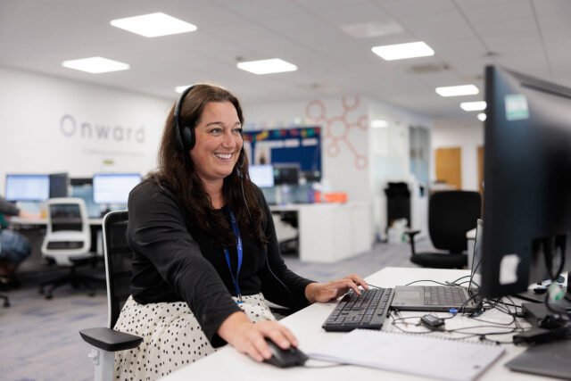 A smiling customer service adviser wearing a headset at a computer desk in the Onward office.