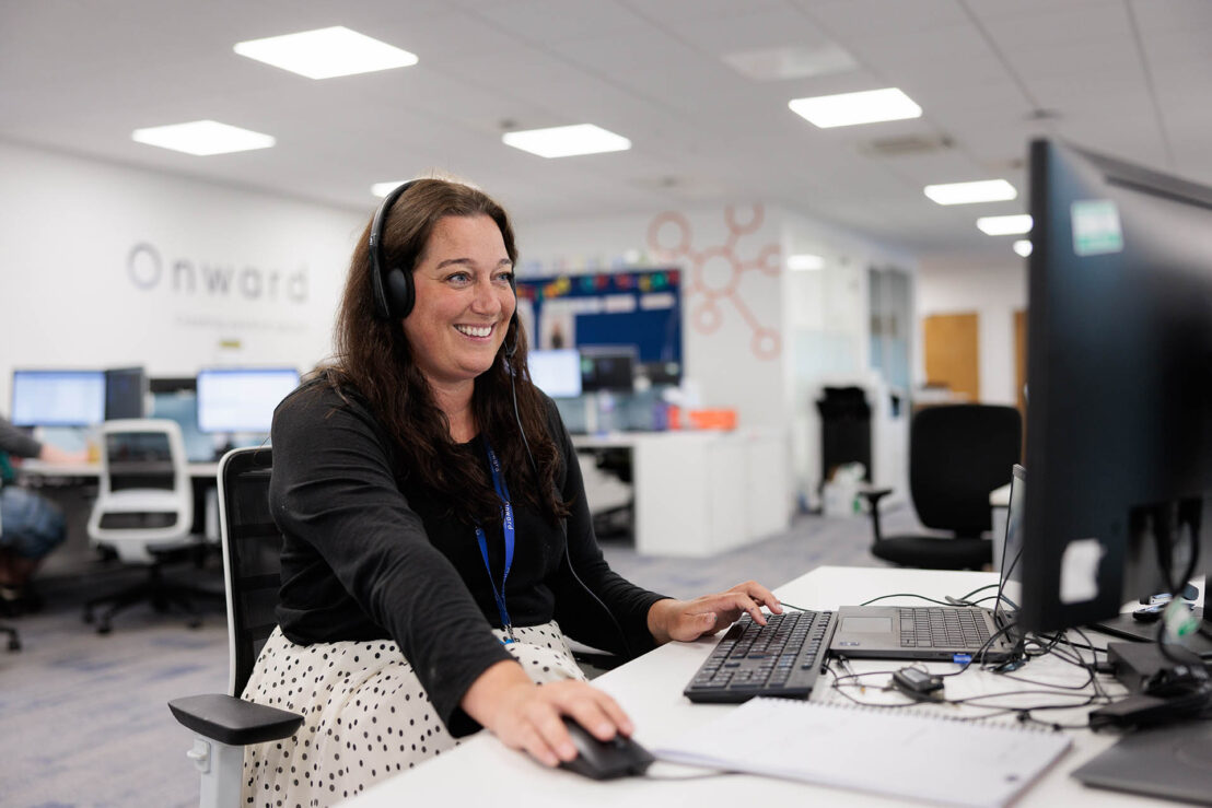 Photograph of a woman wearing a headset working at a computer desk in an office environment. The office features multiple desks with monitors, white walls, and a visible Onward logo on the wall.