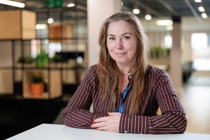 Photograph of a person sitting at a white table in a modern office setting with blurred background featuring plants and shelves. The person wears a maroon striped shirt and a blue Onward lanyard, with hands folded on the table.