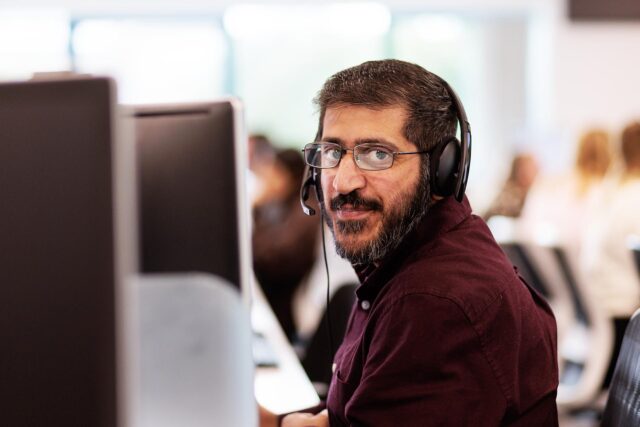 Photograph showing a person wearing a headset and working at a computer in an office environment. Background includes blurred coworkers and natural light from windows, indicating a busy, collaborative workspace.