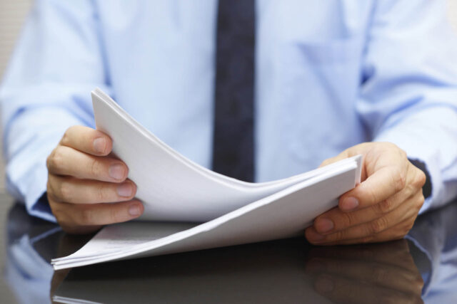 Photograph of a person wearing a blue shirt and dark tie flipping through white papers on a reflective black surface. Focus is on hands and papers, suggesting review or examination of documents.