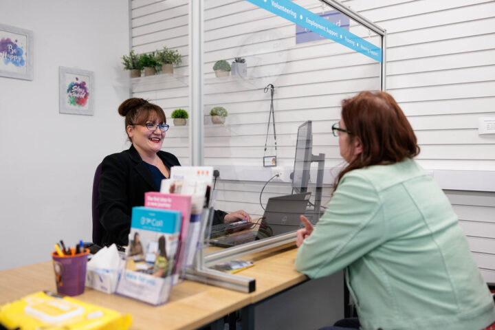 Photograph of two women having a conversation across a desk separated by a clear protective barrier in an office setting. Desk contains brochures, pens, and tissues, while wall behind has small plants and motivational signs.