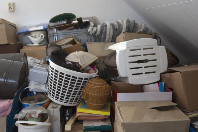 Photograph of a space filled with various household items including cardboard boxes, a white plastic laundry basket, a wicker basket, and folded blankets.