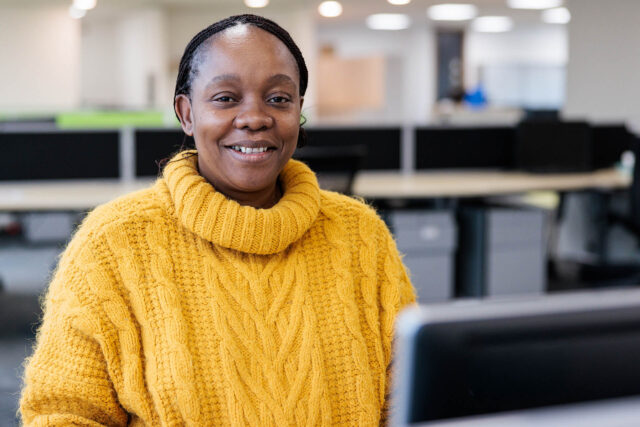 Photograph of a person wearing a yellow cable-knit sweater sitting at a desk in an office environment. Background includes blurred office furniture and another person working.