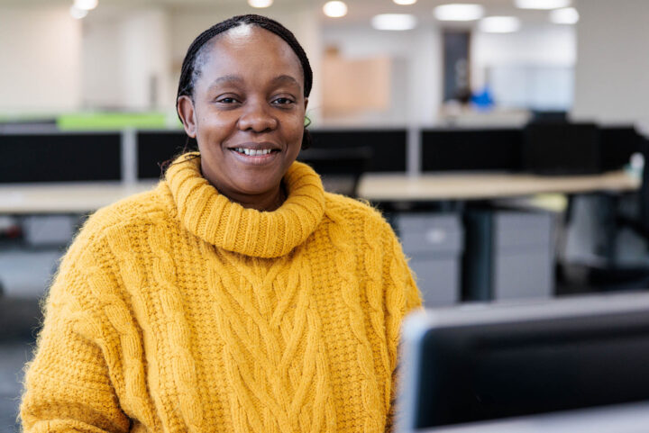Photograph of a person wearing a yellow cable-knit sweater sitting at a desk in an office environment. Background includes blurred office furniture and another person working.