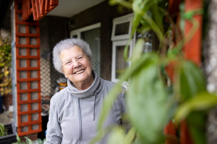Photograph of an elderly person standing outdoors near a building with plants in the foreground. The person wears a light grey hoodie and has short white hair, with a red lattice structure visible on the left side.