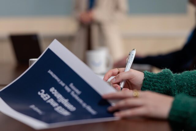 Photograph showing a person holding a pen and reviewing a document. The setting appears to be a meeting or presentation, with blurred figures and a laptop in the background.