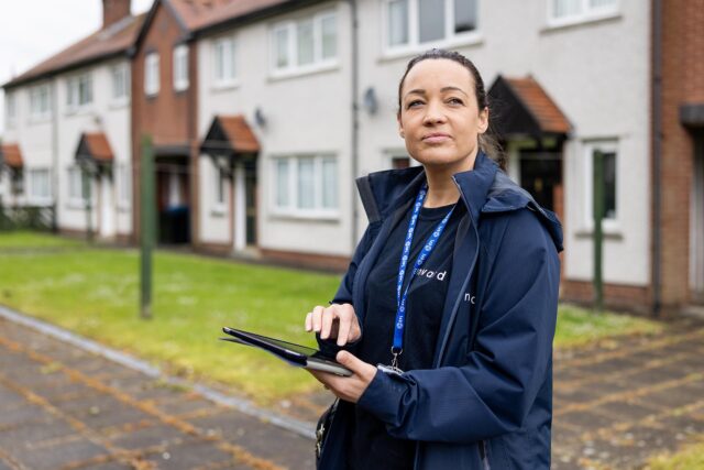 Photograph of a person wearing an Onward jacket and lanyard using a tablet outdoors with houses in the background. The scene suggests data collection or inspection activity.