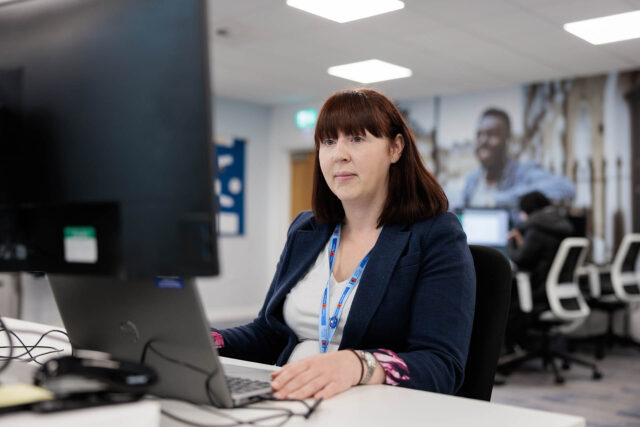 Photograph of a woman working at a computer desk in an office setting with other employees visible in the background.