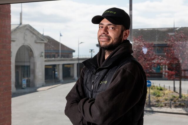An Onward Homes worker in a branded cap and vest standing with arms crossed by a window.