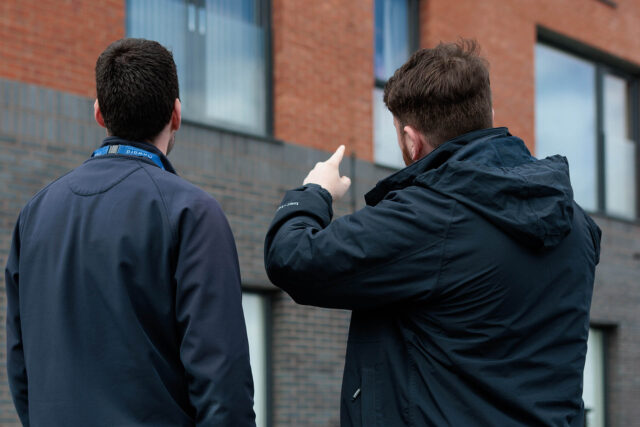 Photograph of two men standing outside a brick and grey stone building, viewed from behind. One man is pointing upward toward the building's windows, suggesting a discussion or inspection.