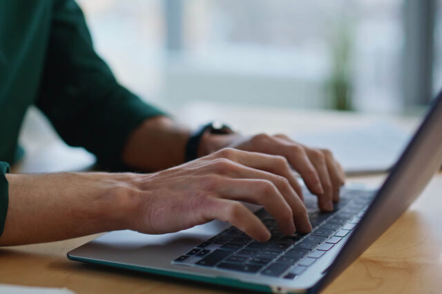 Photograph showing close-up of hands typing on laptop keyboard. Background is blurred with natural light, emphasising focus on hands and device.