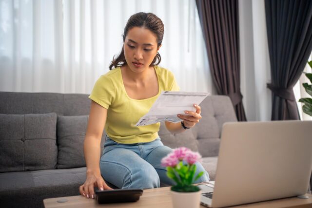 Photograph of a woman sitting on a grey couch holding papers and using a calculator, with a laptop open on a wooden table in front of her. The setting suggests she is managing finances or bills, with natural light coming through white curtains and a small pink flower plant on the table.
