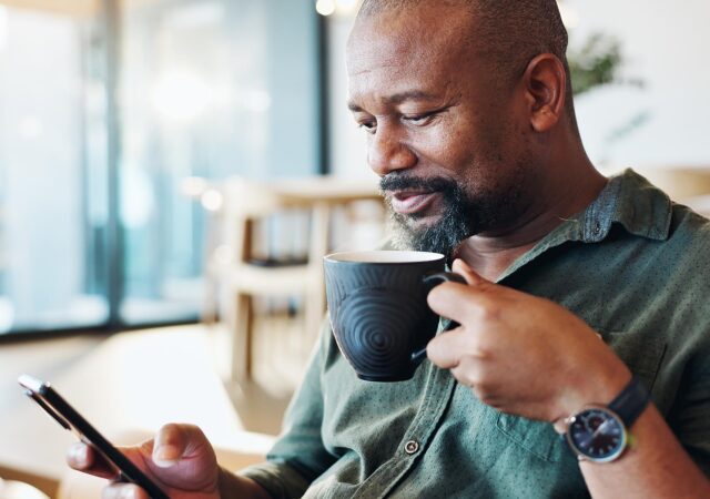Photograph of a person sitting indoors holding a black mug in one hand and a smartphone in the other. The person wears a green button-up shirt and a wristwatch, with a blurred background featuring large windows and furniture.