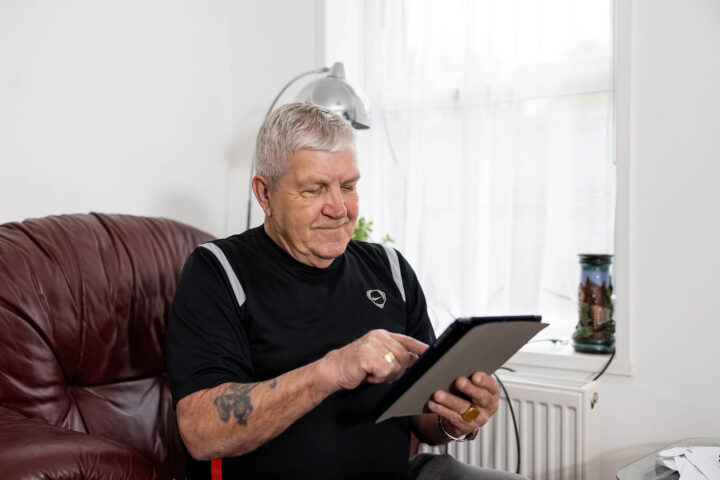 Photograph of an older man sitting on a brown leather couch using a tablet, suggesting engagement with digital technology in a home setting. The scene includes a window with sheer curtains, a silver lamp, and a decorative jar on a white radiator, in a casual and comfortable environment.