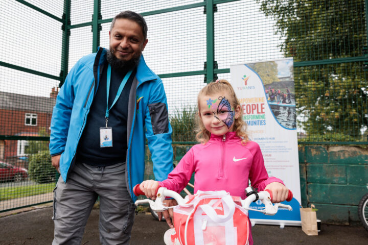 Photograph of a young girl wearing butterfly facepaint on a pink bike facing the camera, smiling. Next to her is a smiling Onward employee, wearing a lanyard and blue jacket, stabilising her bike. They both stand outside, in front of a tall fence.