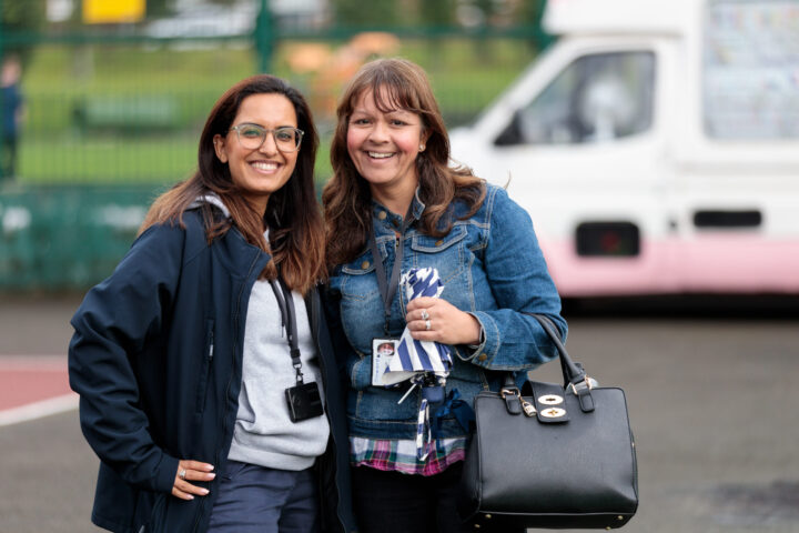 Two women wearing lanyards and casual clothing stand outside in front of a park and ice cream van, facing and smiling at the camera.