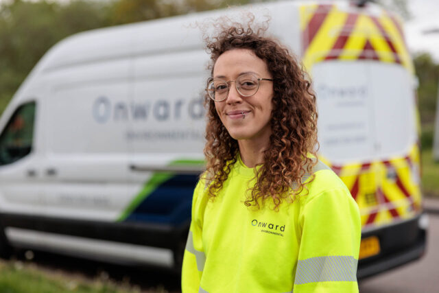 Photograph showing a person wearing a bright yellow high-visibility Onward International sweatshirt standing in front of a white Onward International van with blue and green accents and red-yellow chevron markings on the rear. The image highlights branding on both the clothing and vehicle, suggesting a work or service context related to the company.