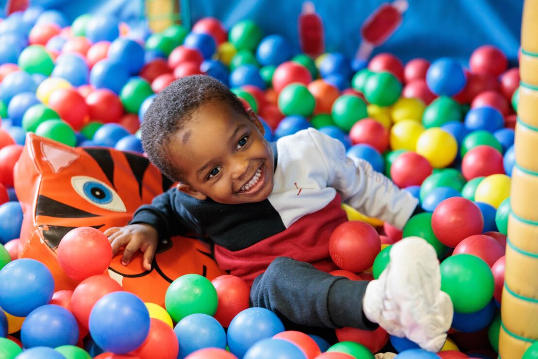 Photograph of a young child playing in a colourful indoor ball pit, smiling while leaning on a soft toy surrounded by red, blue, green, and yellow plastic balls.
