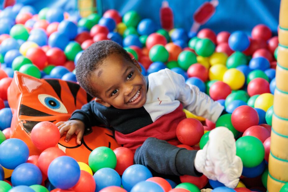 Photograph of a young child playing in a colourful indoor ball pit, smiling while leaning on a soft toy surrounded by red, blue, green, and yellow plastic balls.