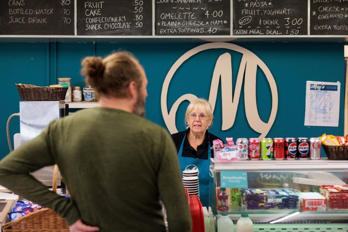 Photograph of a customer standing at a deli counter with a staff member behind it, featuring a large circular logo with a stylized "M" on a teal wall. The counter displays various canned drinks, snacks, and a glass case with packaged food, while a blackboard menu above lists items and prices in white chalk.