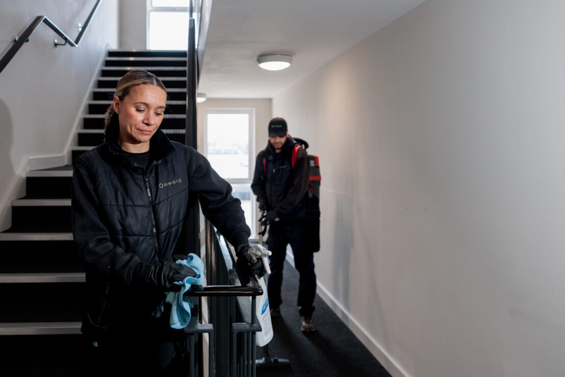 Photograph showing two people cleaning a stairwell and hallway in a building. One person is wiping a handrail with a blue cloth while the other is vacuuming the carpeted floor, both wearing black uniforms and gloves.