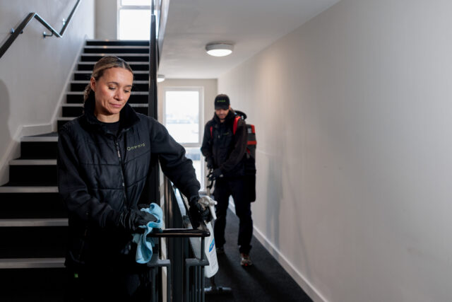 Photograph showing two people cleaning a stairwell and hallway in a building. One person is wiping a handrail with a blue cloth while the other is vacuuming the carpeted floor, both wearing black uniforms and gloves.