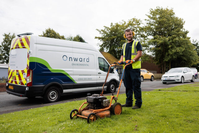 Photograph of a worker wearing a high-visibility vest operating a lawn mower on a grassy area beside a street. A white van with "Onward Environmental" branding and contact information is parked on the road behind the worker, with several cars visible further down the street.