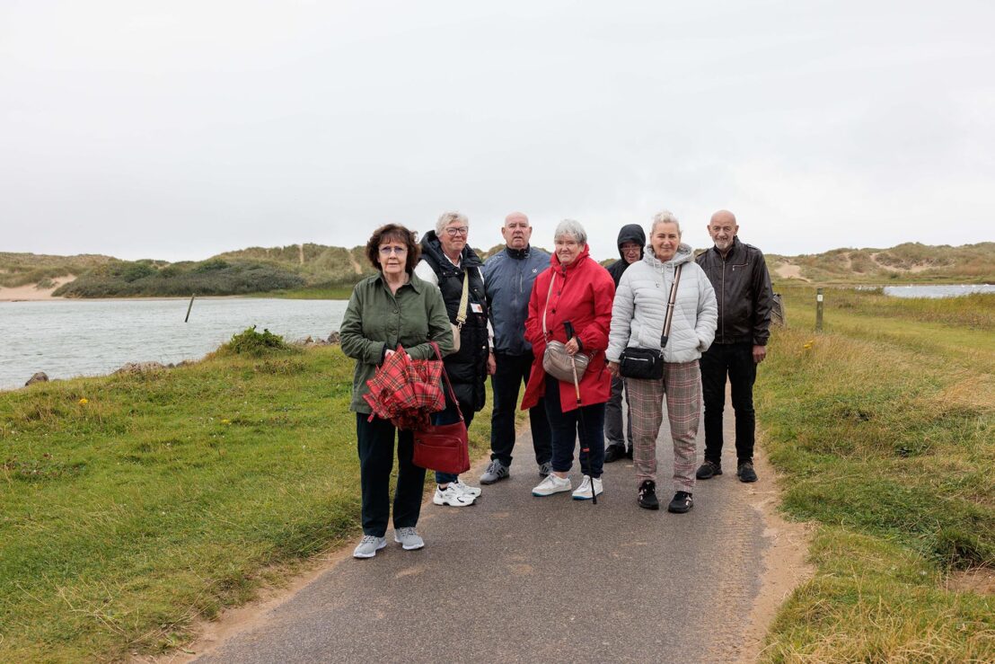 Photograph of a group of seven older adults standing on a paved path near a body of water and grassy dunes under an overcast sky. The group wears casual jackets and carries bags, with one person holding a folded red umbrella, suggesting a leisurely outdoor walk or gathering.