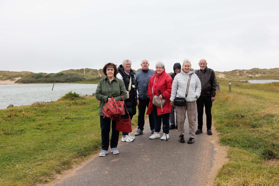 Photograph of a group of seven older adults standing on a paved path near a body of water and grassy dunes under an overcast sky. The group wears casual jackets and carries bags, with one person holding a folded red umbrella, suggesting a leisurely outdoor walk or gathering.