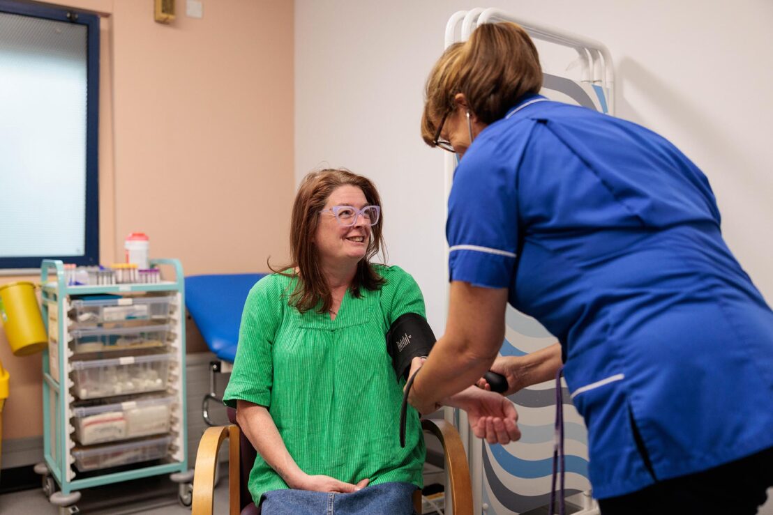 Photograph showing a healthcare professional in blue uniform measuring blood pressure of a seated patient wearing a green shirt. Setting appears to be a medical examination room with medical supplies and equipment visible in background.