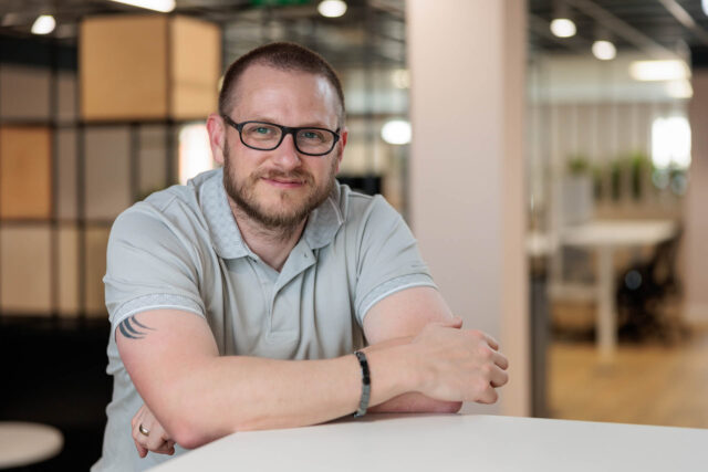 Photograph of a man sitting at a white table in a modern office setting with blurred background.