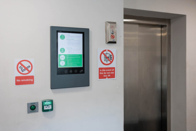 Photograph of a wall next to an elevator door featuring safety and informational signage. Signs include a red "No smoking" symbol, an "In the event of fire do not use this lift" warning, a green emergency exit button, and a digital display panel with instructions or information.