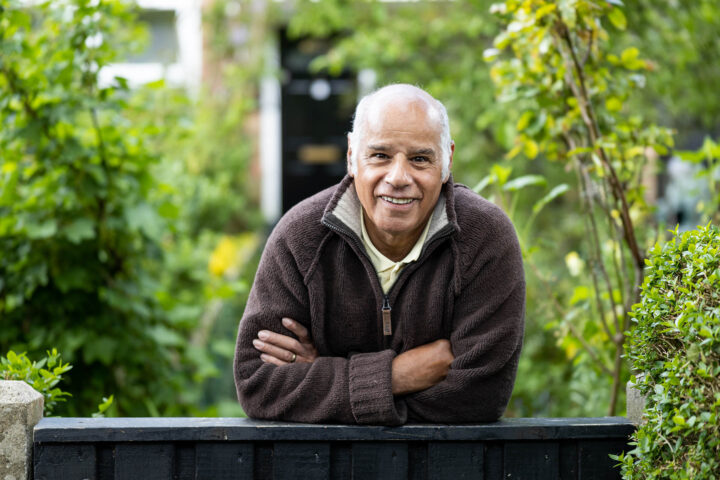 Photograph of an elderly person leaning on a black wooden fence surrounded by lush green plants and trees. The person wears a brown zip-up sweater and a white collared shirt, with a residential building visible in the background.