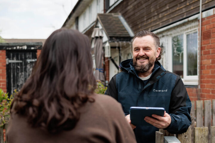 Photograph showing a person wearing a dark jacket with "Onward" logo holding a tablet and speaking to another person outside a residential building. The scene suggests a home visit or inspection, with a focus on interaction between the two individuals.