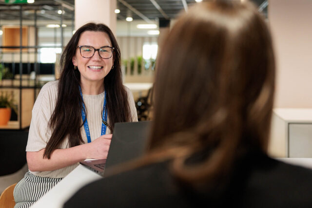 Photograph of two women engaged in a conversation across a desk in a modern office setting, with one woman facing the camera and the other seen from behind. The woman facing the camera wears a white shirt and blue lanyard, using a laptop, suggesting a professional or interview context.