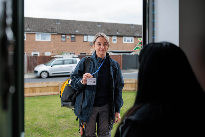 Photograph showing a repairs operative standing outside a doorway holding up an identification badge, wearing a dark jacket and carrying a backpack. A second person is partially visible inside, with a suburban neighbourhood and parked car in the background.
