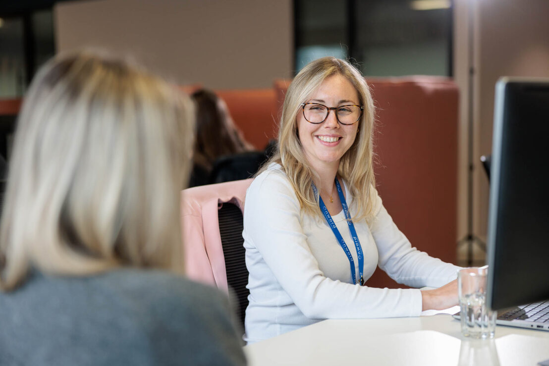 Photograph showing two women in an office setting engaged in conversation, with one woman facing the camera and the other with her back to it. The woman facing the camera wears a white top and blue lanyard, seated at a desk with a computer monitor and glass of water.