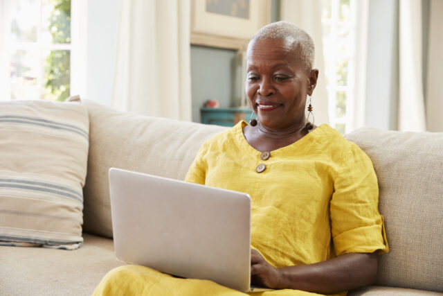 Photograph of a person sitting on a beige couch using a laptop, wearing a bright yellow outfit. Background includes a striped pillow and a window with natural light illuminating the room.