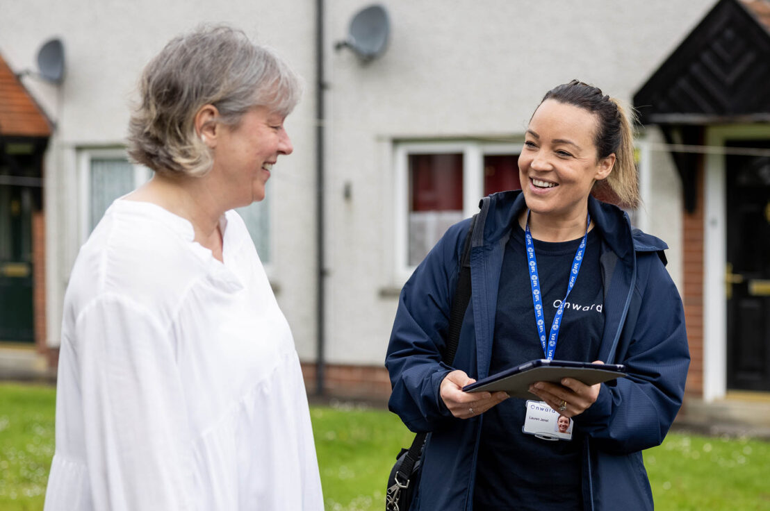Photograph showing a worker wearing a navy jacket and holding a clipboard, engaging in conversation with an elderly woman dressed in white outside a residential building. The worker wears a lanyard with an ID badge, indicating a professional visit or assessment in a community or home care setting.