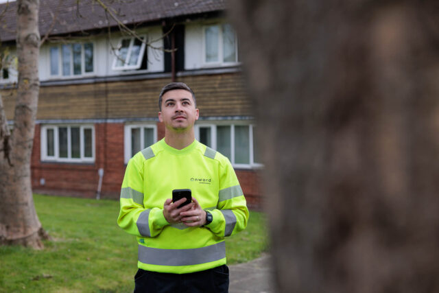 Photograph of a person wearing a bright yellow high-visibility jacket standing outdoors near a residential building while holding a smartphone. The jacket features reflective silver stripes and a logo on the chest, indicating a work or safety-related context.