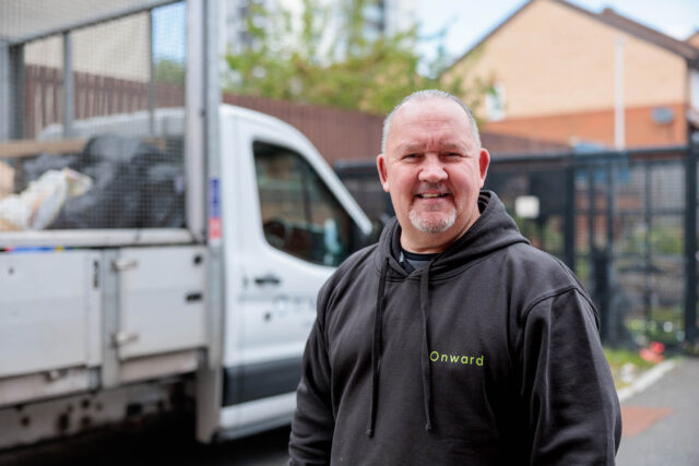Photograph of a person wearing a black hoodie with "Onward" printed on the chest, standing outdoors near a white truck loaded with bags. Background includes residential buildings and a fence, suggesting a work or delivery context.