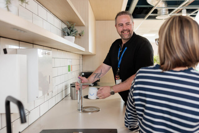 Photograph of two people in a modern kitchen or break room, with one person filling a white mug from a water dispenser on a tiled countertop. The setting includes light wood cabinetry, white tiled backsplash, and a potted plant on a shelf, suggesting an office environment.
