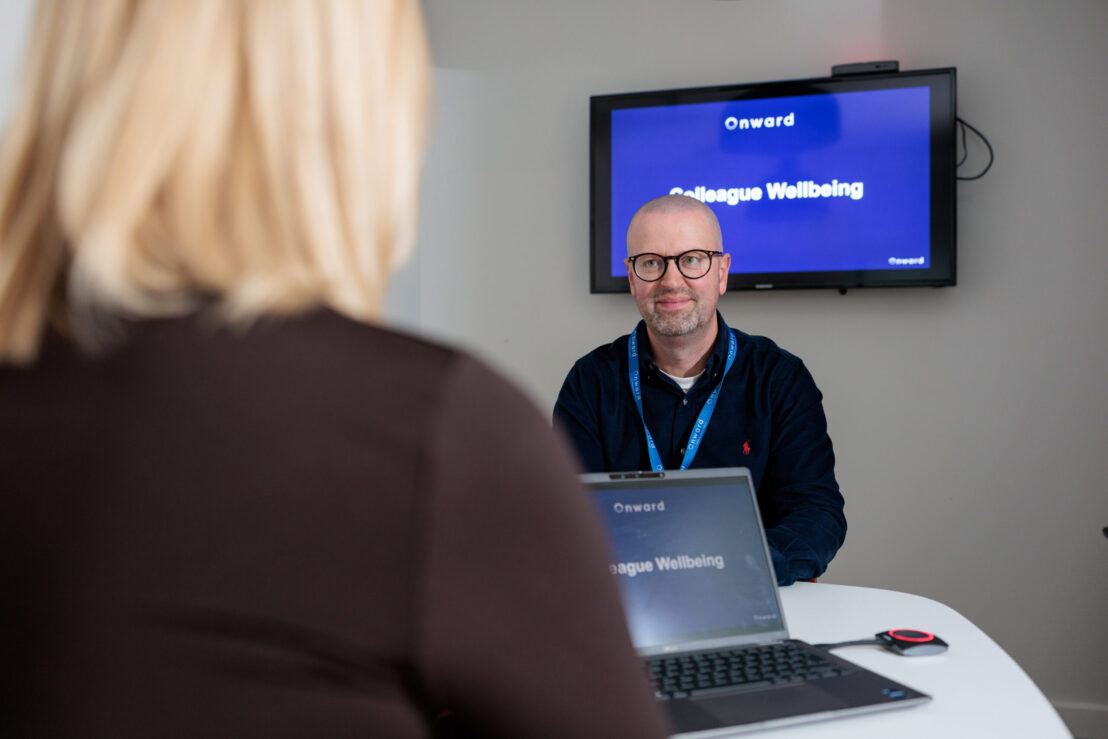Photograph of a man wearing glasses wearing a lanyard with a wall-mounted screen behind him that reads "Onward: colleague wellbeing". In the foreground a woman stands with her back to the camera, in conversation with the man.
