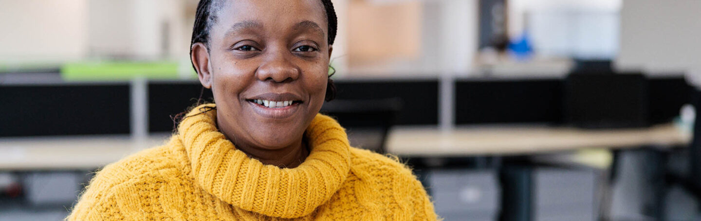 Photograph of a person wearing a yellow cable-knit sweater sitting at a desk in an office environment. Background includes blurred office furniture and another person working.